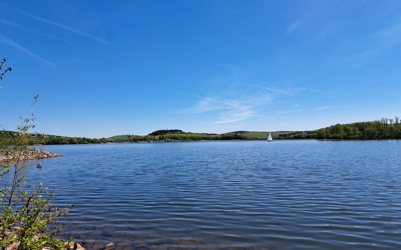 Es ist ein See zu sehen mit grün bewaldetem Ufer. Auf dem See ist ein Segelschiff.